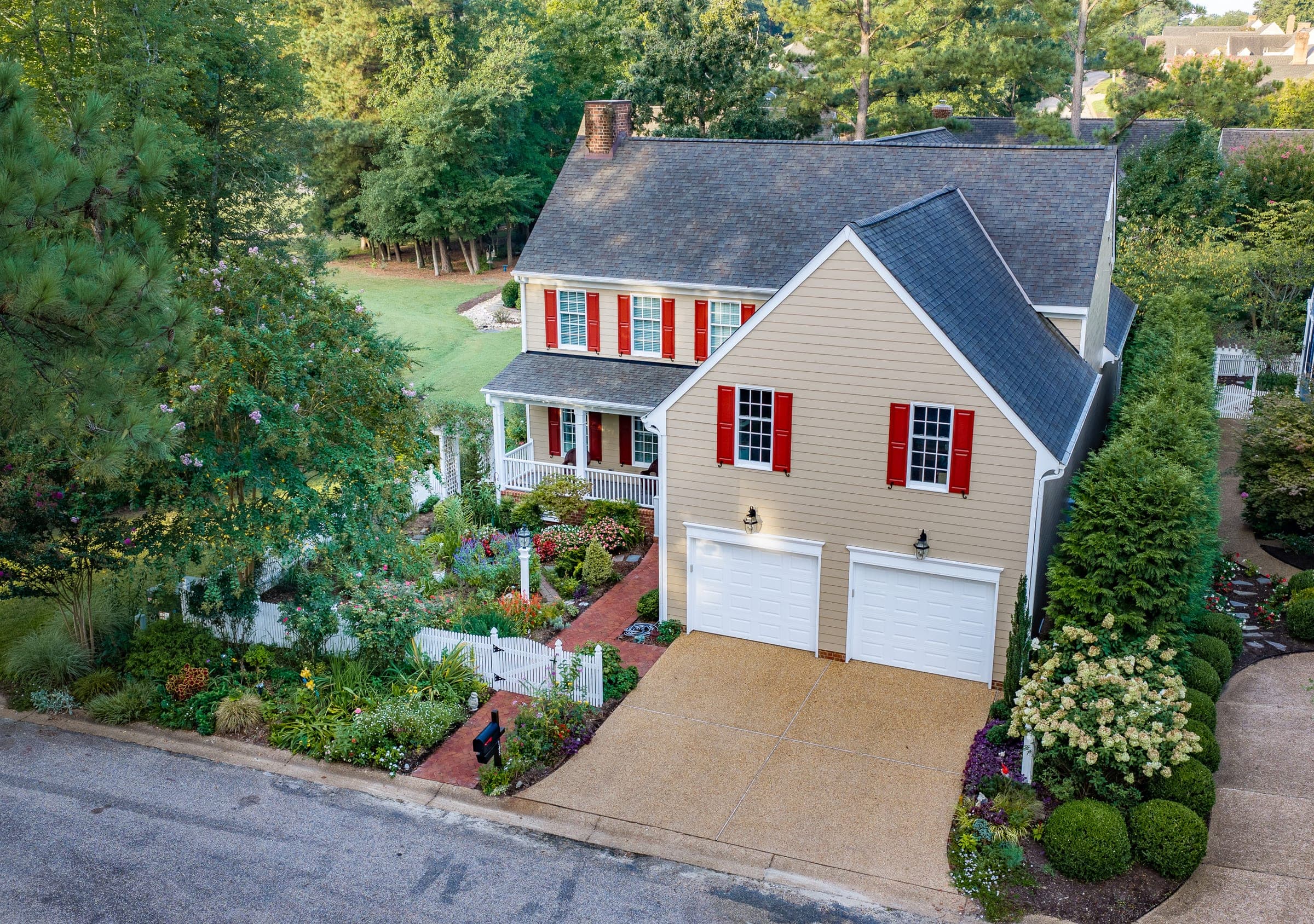 145 Hollinwell: aerial view of a tan two-story home with red shutters, a white picket fence, brick walks, and layered gardens in Ford's Colony